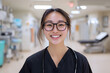 © Wittawit - smiling female doctor in medical facility, wearing glasses and stethoscope, exuding confidence and warmth. background shows medical equipment and treatment area