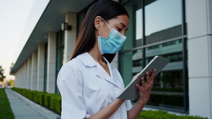 Poster - Female healthcare professional in surgical mask and white coat analyzing medical data on tablet computer, standing outside modern hospital building in slow motion montage sequence