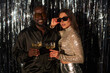 © DragonImages - Smiling couple enjoying festive celebration and holding champagne glasses in stylish outfits while standing against shimmering background