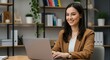 © PP Creative - A professional and young businesswoman with long dark hair and a warm smile working on her laptop in a modern office setting