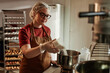© Seventyfour - Medium shot of young female bakehouse worker adding shredded coconut into bowl precisely measuring ingredient on scales of while cooking in bakery kitchen