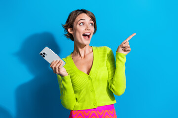 Aufkleber - Photo of young surprised lady in green shirt direct finger empty space holding smartphone online store isolated on blue color background