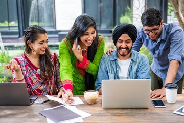 Poster - Group of four people collaborating at a table with laptops and notebooks, smiling and engaged in teamwork, outdoors in a casual setting. Indian students working together on laptop.