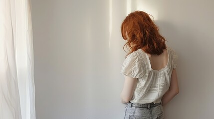 Wall Mural - Young woman with red hair stands against a white wall, showcasing a serene moment in a softly lit room