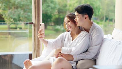 Wall Mural - Asia people young adult man woman relax smile look camera take photo post to social media vlog video at outdoor hotel balcony enjoy date day. Happy asian lover wife husband just married sweet newlywed