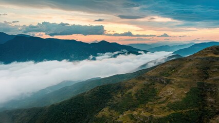 Wall Mural - Majestic mountain landscape with gentle clouds at sunset, deep greens in foreground, layered blues in background, creating a serene vista.