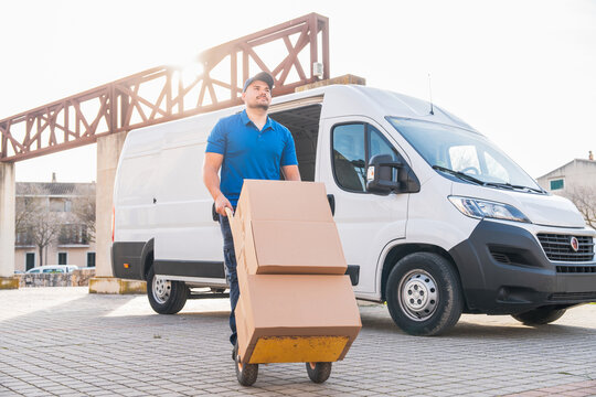 Delivery driver transporting packages with hand truck in front of van. Delivery driver concept in a van. Man in blue uniform moving boxes with a hand truck, representing logistics and distribution.