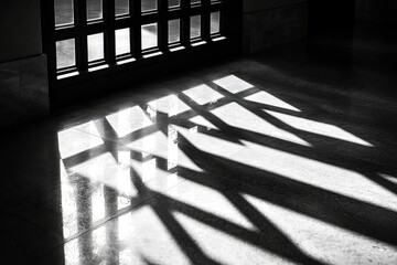 Poster - A grayscale capture of sunlight streaming through a multi-paned window, casting intricate patterns of light and shadow onto the tiled floor of an empty interior.