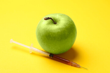 Wall Mural - GMO concept. Green apple and syringe with liquid on yellow background, closeup