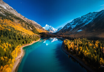 Poster - Aerial view of a stunning mountain valley with a turquoise lake during autumn. Colorful trees on slopes and snowy peaks under a clear blue sky.