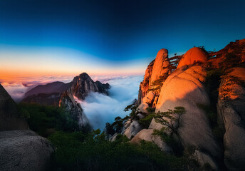 Poster - Mountain peaks pierce through clouds at sunset. Golden light bathes rocky slopes as the sky blends blue and orange hues. A scenic, tranquil vista.