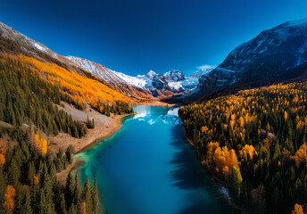 Poster - An aerial view shows a vibrant autumn valley with a blue lake nestled between colorful forests and snow-capped mountains under a clear sky.