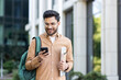 © Liubomir - Young student with backpack on shoulder using app on smartphone. Man walking outside university campus smiling with phone in hands.