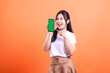 © Ajab - A woman with long dark hair is holding a smart phone device with a green screen and pointing at it with her index finger, isolated on orange background.