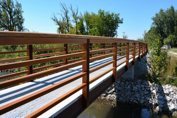 Aufkleber - Serene pedestrian bridge over calm stream with lush greenery and clear blue sky in outdoor setting