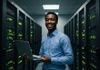 © ferifadli - A young African American man carrying a laptop in a server room, smiling at the camera
