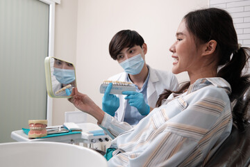 Wall Mural - Asian male dentist explains color shades of tooth implant whitening to young female patient, look at mirror in dental clinic, and do professional mouth hygiene healthcare work in a doctor's hospital.