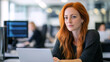 © ibooM - A woman with red hair in casual attire sits at a laptop in a modern office. She displays concentration and professionalism in a dynamic workspace filled with technology.