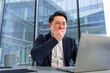 © RomArt production - Professional Asian businessman in a modern office startled while looking at a laptop. Depicts surprise, work life, and business challenges in a corporate setting. Business-casual attire urban context.