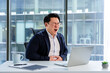 © RomArt production - Sick Asian businessman sitting in a modern office space, feeling discomfort while looking at a laptop. Stylishly dressed in business attire, the workspace includes bright lighting and office items.