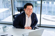 © RomArt production - Asian businessman seated in a modern office appears uncomfortable, portraying moments of distress. Wearing formal attire, the image exudes emotions of tension and corporate work-related stress.