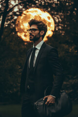 fashionable man in a tailored suit holding a bag, posed in front of a dramatic full moon and tree ba