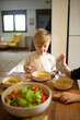 © Mariia Andreeva - A young boy enjoys a healthy meal with his family, eating soup and salad with great appetite. The scene reflects wholesome nutrition, warmth, and togetherness at the dining table.