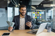 © Liubomir - Successful and happy businessman sitting at workplace inside office, man holding cash money dollars in hands. Man in business suit using app on smartphone