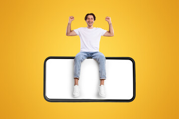 Poster - Yes. Excited Man Sitting On Big Smartphone With Empty White Screen Shaking Clenched Fists, Cheerful Handsome Guy Celebrating Win Isolated On Yellow Orange Background, Mock Up Collage, Banner Panorama