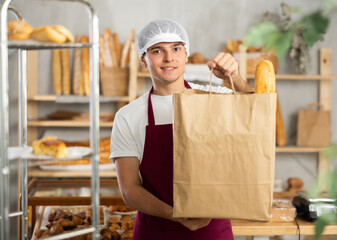 Portrait of positive young baker with paper bag of fresh baked goods in the interior of a private bakery