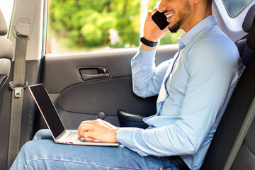 Wall Mural - Wealthy middle-eastern businessman sitting in car, using laptop and having business conversation on smartphone, working online while going to airport, copy space. Modern technologies in business