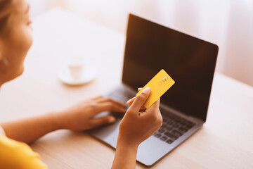 Poster - Online Shopping Concept. Over the shoulder of african american woman holding credit card in hand and using laptop with black blank screen, typing on keyboard. Selective focus, blurred background