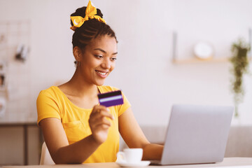 Poster - Online Shopping Concept. Portrait of smiling african american woman holding credit card in hand and using laptop, sitting at desk, typing on keyboard, blurred background, selective focus. Ecommerce