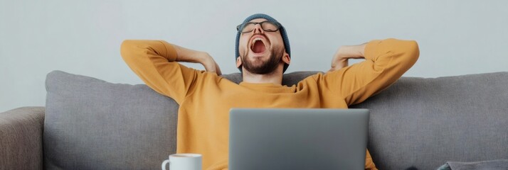 Poster - Tired man wearing glasses and a beanie yawns while sitting on a couch with a laptop, indicating stress and fatigue from multitasking and managing deadlines in a remote work environment