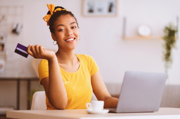 Poster - Online Shopping Concept. Portrait of smiling african american woman holding credit card in hand and using laptop, ordering gifts, typing on keyboard, looking at camera, blurred background