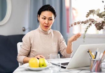 Wall Mural - Friendly asian woman sitting at a table in an apartment, communicates with someone via video link on a laptop