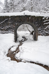 Leinwandbilder - Snow covered landscape featuring an arch bridge over a small stream in a winter wonderland setting