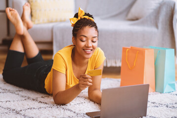 Poster - Online Transaction Concept. Smiling black lady using laptop and credit card, ordering purchases via internet, lying on the floor carpet, checking debit card number and typing cvv security code