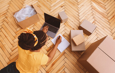 Poster - Above top high angle view of young african american woman typing on laptop keyboard, writing in notebook, lying on the floor near cardboard boxes. Computer with blank empty screen for mockup
