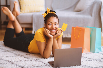 Poster - Online Shopping Concept. Portrait of cute black young lady lying on the floor carpet at home with computer, holding debit credit card and looking at camera, ordering clothes on internet