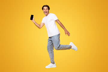 Poster - Happy young man showing empty smartphone screen while jumping up over yellow studio background, full body length shot. Excited running guy recommending nice and useful mobile application