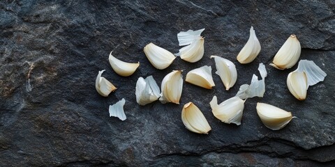 Poster - Garlic cloves scattered on dark textured stone background with light and shadow, showcasing their natural color and shape for culinary use.