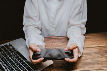 Poster - Woman Holding Smartphone Above Wooden Desk with Laptop in Background