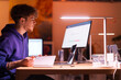 © Studio Marmellata - A young man in a purple hoodie sits at a desk in a warmly lit office, focused on his laptop while holding a pencil, with a monitor displaying financial charts nearby.