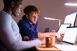 © Studio Marmellata - Two young professionals sit at a shared desk in a dimly lit office, reviewing stock market data displayed on multiple screens while discussing investment strategies.