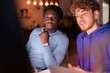 © Studio Marmellata - Two young men work together at a desk in a modern office, reviewing data on multiple screens while collaborating on a financial or business-related project.