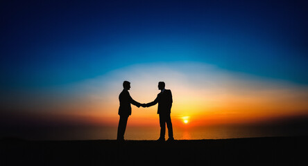 Aufkleber - Two silhouetted figures shaking hands against a vibrant sunset sky. The horizon glows orange and yellow, contrasting with the dark figures and the deep blue sky above.