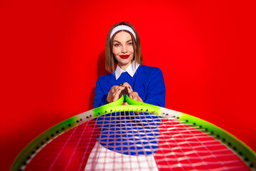 Stylish woman posing with a tennis racket against vibrant red background, wearing a blue jumper and headband