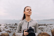 © SHOTPRIME STUDIO - Young woman with a camera at the beach, smiling joyfully while embracing her passion for photography, wearing a gray sports hoodie in a coastal setting