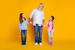 © deagreez - Grandfather holding hands with his two smiling granddaughters against a vibrant yellow background, wearing casual clothing, generating a feeling of family connection and joyful bonding.